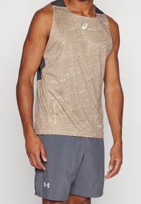 Beżowa, bezrękawnikowa koszulka sportowa z teksturowanym wzorem; szare akcenty na ramionach. W połączeniu z szarymi szortami z logo Under Armour.