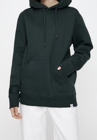 Personne portant un sweat à capuche vert foncé avec une poche avant et un pantalon blanc, une main dans la poche et l'autre détendue le long du corps.