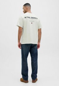 Lichtgrijze T-shirt met "ROYAL DENIM" in zwart gedrukt op de achterkant, gecombineerd met donkerblauwe jeans en bruine schoenen. Casual pasvorm.
