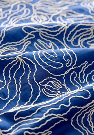 Blått tyg med vitt broderat spiralmönster, med mjuk textur och slät yta, som framhäver intrikata, flödande linjer.