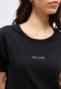 Γυναίκα που φοράει μαύρο t-shirt με στρογγυλή λαιμόκοψη και λογότυπο "PEPE JEANS" από μικρές λευκές κουκκίδες στο στήθος.
