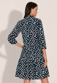 Dunkelblaues Kleid mit weißem Punkte-Muster, ¾-Ärmel und einem gestuften Volant-Saum. Hergestellt aus einem glatten Stoff mit fließender Silhouette.