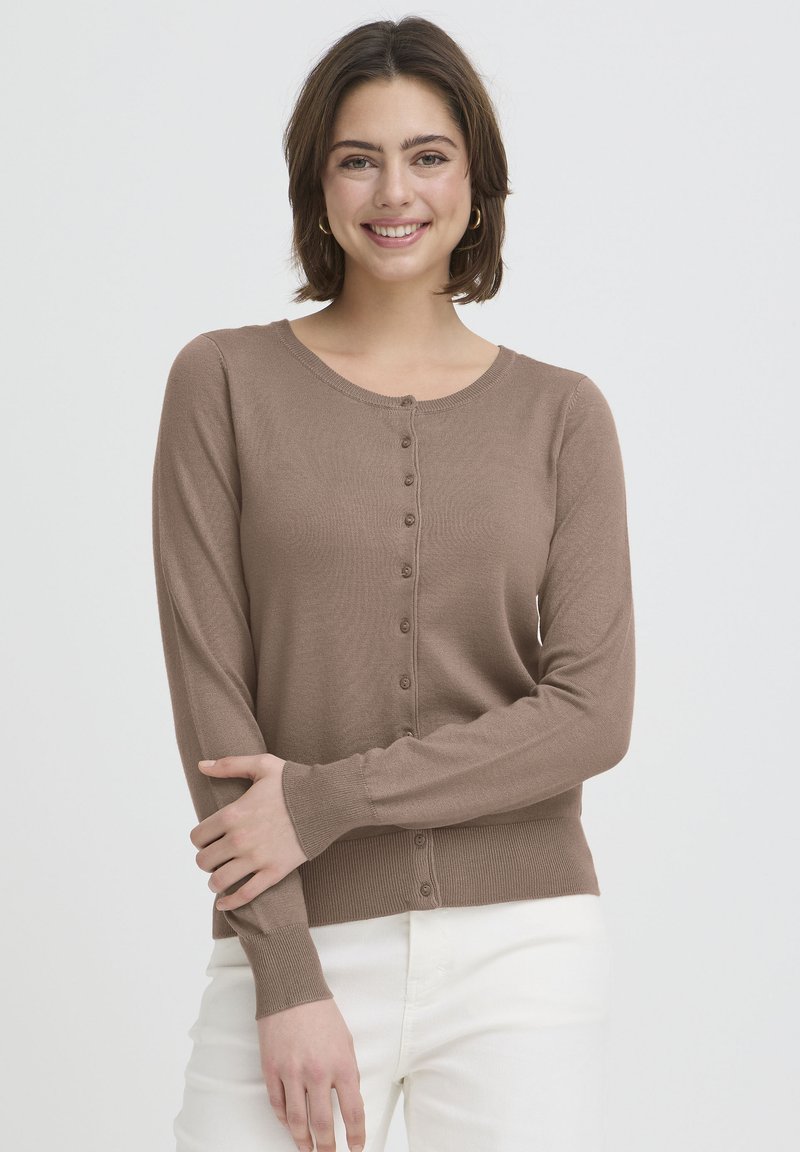 Cardigan marron à manches longues en tissu côtelé avec un col rond et sept boutons sur le devant, associé à un pantalon blanc.