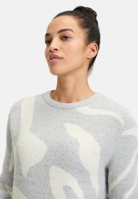 Hellgrauer Pullover aus strukturiertem Strick, mit abstrakten weißen Mustern und einem klassischen Rundhalsausschnitt. Weiches, gemütliches Material.