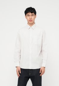 Νέος άνδρας που κοιτάζει μπροστά, φοράει λευκό πουκάμισο με μακριά μανίκια και λεπτές κάθετες ρίγες και σκούρο τζιν, σε απλό φόντο.