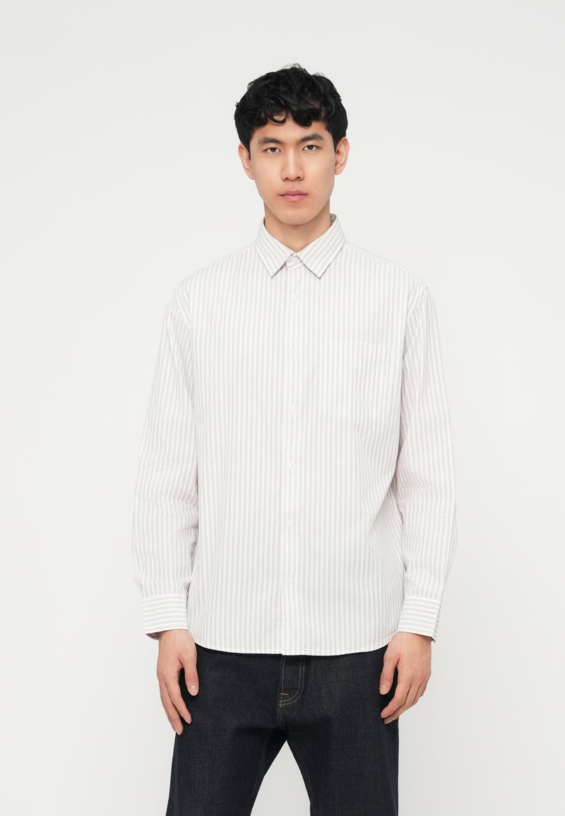 Νέος άνδρας που κοιτάζει μπροστά, φοράει λευκό πουκάμισο με μακριά μανίκια και λεπτές κάθετες ρίγες και σκούρο τζιν, σε απλό φόντο.