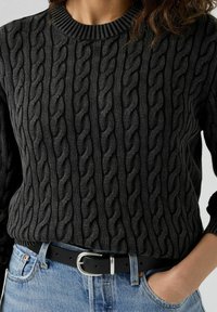 Svart kabelstickad tröja med rund halsringning, strukturerat mönster och ribbade ärmslut, tillsammans med blå denim och ett svart bälte med en silverspänne.