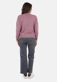 Donna in piedi di spalle, indossa un maglione a collo alto color malva, jeans dritti grigi e sneakers bianche con dettagli neri sul tallone.