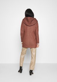 Abrigo de lana de color óxido con capucha, con un corte entallado y costuras visibles. Llevado sobre pantalones de cuero sintético beige y botas de tacón negras.