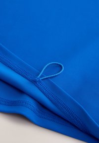 Lysende blåt stof med en glat tekstur, med en løkke-detalje og syede kanter for holdbarhed. Blødt og fleksibelt materiale.