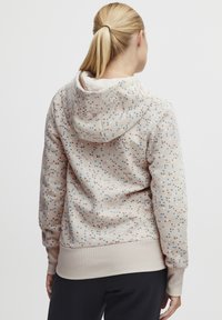 Hettegenser i myk beige stoff med en teksturert overflate, med et flerfarget prikkemønster i rødt, blått og brunt. Ribbestrikkede mansjetter.