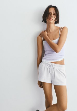 Mladi žena s kratkom tamnom kosom u bijelom topu i svijetlosivim kratkim hlačama s vezicama stoji nasuprot običnog bijelog zida.