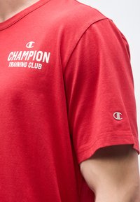 Κόκκινο πουκάμισο με κοντά μανίκια, με λευκό κείμενο "Champion Training Club" στο στήθος και μικρό λογότυπο Champion στο μανίκι, φορεμένο από ένα άτομο.