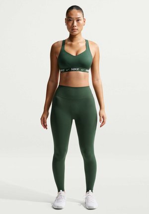 Femme debout, face à l'avant, portant un brassard de sport Nike vert foncé et un legging assorti, avec des baskets blanches, sur un fond clair uni.