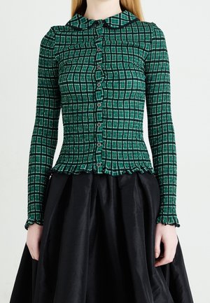 Donna con una camicia aderente a quadri verdi con bordi arricciati e una gonna nera, in piedi davanti a uno sfondo bianco uniforme.