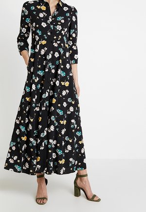 Kvinde iført en sort lang blomsterkjole med grønne, hvide og gule blomster, kombineret med olivengrønne højhælede sandaler.