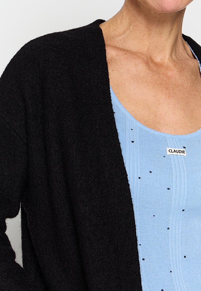 Schwarze, weiche Strickjacke mit offenem Vorderteil, getragen über einem hellblauen Stricktop mit schwarzen Sprenkeln und einem Etikett mit der Aufschrift "CLAUDIE."