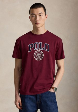 Młody mężczyzna o krótkich włosach, ubrany w bordową koszulkę Polo Ralph Lauren i niebieskie dżinsy, stojący na jednolitym beżowym tle.