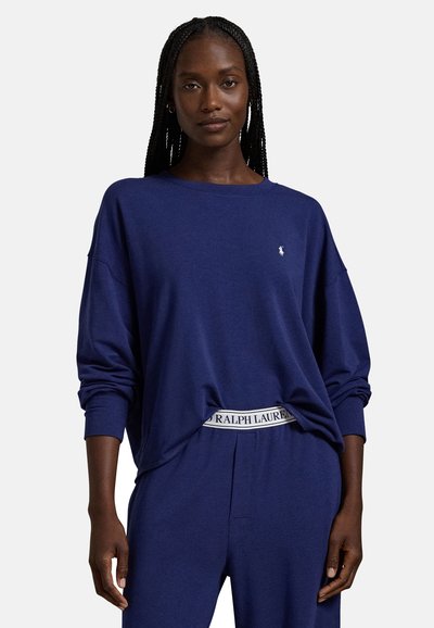 Γυναίκα που φοράει μπλε φουτερ με μακριά μανίκια και παντελόνι με το ίδιο χρώμα, με ορατή λευκή ζώνη Ralph Lauren, στέκεται μπροστά σε απλό φόντο.