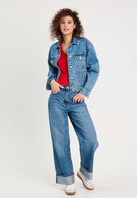 Jeansjacke in hellblau, mit Knopfleiste und Brusttaschen. Kombiniert mit lockeren blauen Jeans und weißen Turnschuhen mit braunen Sohlen.