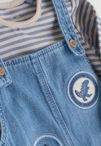 Pantalones de mezclilla azul para bebé con botones de madera y un parche redondo que muestra un dinosaurio azul con el texto "Increíble pequeño".