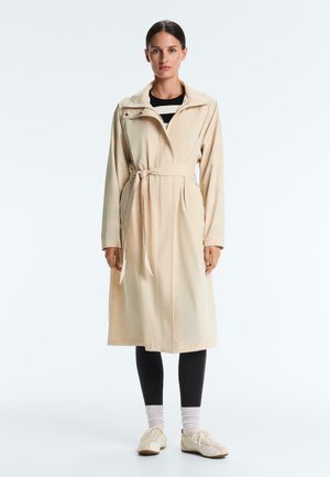 Femeie purtând o trenci bej cu curea peste un top cu dungi alb-negru, colanți negri, șosete albe și adidași bej, stând în poziție verticală.