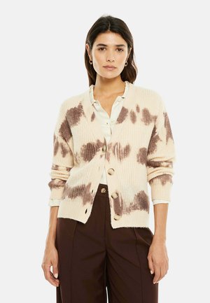 Mujer con cárdigan crema con manchas marrones de teñido anudado, blusa crema y pantalones anchos de tiro alto en marrón oscuro, de pie sobre fondo blanco.