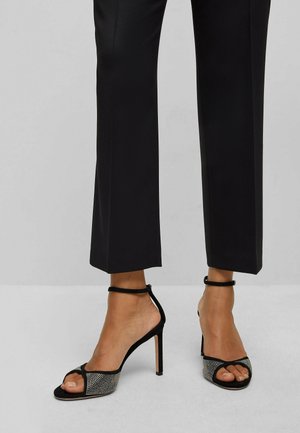 Czarne sandały na wysokim obcasie mają otwarty nos z błyszczącym, zdobionym przodem oraz paskiem na kostce. Materiał wydaje się miękki i o teksturze.