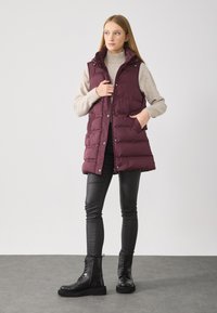 Burgunderfarbene Steppweste mit Druckknöpfen und seitlichen Taschen, getragen über einem beigen Pullover. Kombiniert mit schwarzen Slim-Fit-Hosen und Schnürstiefeln.