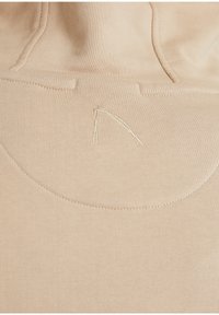 Primo piano di un tessuto beige con un sottile logo triangolare cucito e dettagli delle cuciture su un indumento.