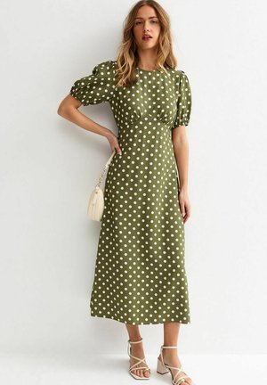 Kobieta w oliwkowo-zielonej sukience do połowy łydki w białe grochy, z bufkami na rękawach, trzymająca beżową torebkę, nosząca białe sandały na szerokim obcasie z paskami.