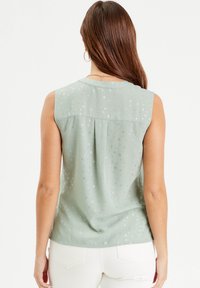 Chemisier sans manches en tissu vert clair avec un motif floral et des accents métalliques, doté d'un pli au dos pour plus de détail et d'aisance.