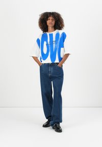 White oversized t-shirt with blue abstract pattern and black text. Paired with loose dark blue jeans and black shoes. Plain background.