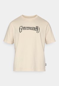 Μπεζ βαμβακερό t-shirt με μαύρο γραφικό "GRATEFULDEAD" στο στήθος, κοντά μανίκια και άνετη εφαρμογή. Ετικέτα ορατή στην πλευρά.