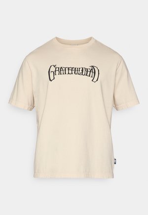 Μπεζ βαμβακερό t-shirt με μαύρο γραφικό "GRATEFULDEAD" στο στήθος, κοντά μανίκια και άνετη εφαρμογή. Ετικέτα ορατή στην πλευρά.