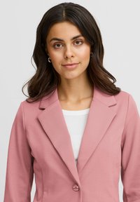 Roze tailleurjasje met notch-revers, enkele knoopsluiting en een gladde textuur, gedragen over een witte aansluitende top.