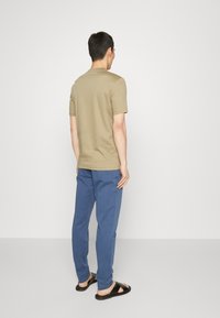 Chemise à manches courtes vert olive associée à un pantalon bleu. La chemise présente un col rond, et la tenue est complétée par des sandales noires.