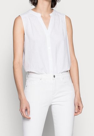 Femme portant un chemisier blanc sans manches à boutons et un pantalon blanc taille haute sur un fond uni.