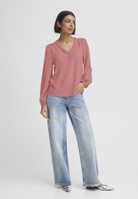 Langærmet bluse med V-hals i støvet rosa, kombineret med lyseblå jeans med vide ben. Modellen har sølvfarvede spidse sko på. Glat tekstur.
