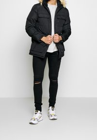 Chaqueta de abrigo negra con capucha forrada de pelo, bolsillos frontales y cierre de cremallera. Lleva puesta una camiseta gris claro y está combinada con jeans rotos negros y zapatillas.