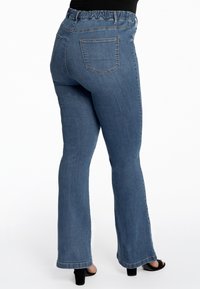 Jean en denim évasés bleu moyen, présentant une taille élastique, des poches arrière et une texture lisse avec des accents de couture sur l'ourlet.