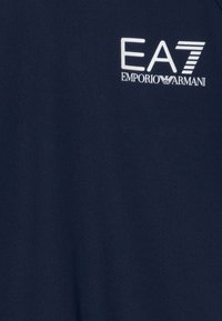 Polo en bleu marine en tissu texturé. Présente un logo blanc : "EA7 EMPORIO ARMANI." Design simple, manches courtes et col classique.