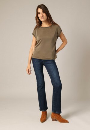 Olijfgroene top met korte mouwen en een gladde textuur; gecombineerd met donkerblauwe recht gesneden jeans en bruin suède enkellaarsjes.