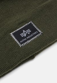 Olivovo zelená pletená čiapka s čiernou nášivkou a bielym stehom, na ktorej je emblém. Materiál vykazuje jemnú textúru a pružnosť.