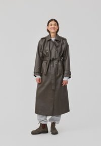 Lang mørkegrønn lær trench coat med dobbeltknapper, belte i midjen og skulderklaffer, kombinert med grå joggebukser og brune støvler.