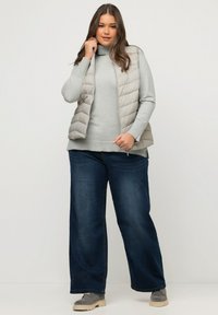 Beżowa kamizelka puchowa nałożona na jasnoszary sweter, połączona z szerokimi dżinsami w ciemnym odcieniu. Kamizelka ma zamek błyskawiczny i boczne kieszenie. Szare obuwie.
