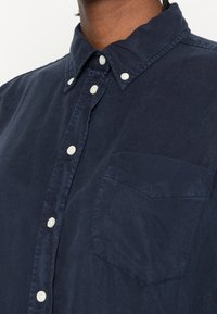 Navyblaues Button-Up-Hemd aus Baumwolle, mit klassischem Kragen, weißen Knöpfen und einer einzelnen Brusttasche im flachen Design.
