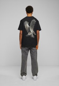 Sort t-shirt med et stort ørne-grafik på ryggen, med grå denimbukser og hvide sneakers. Orange mærke nær kanten.