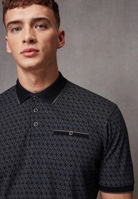Polo-shirt bleumarin cu un design patternat, având o guler solid negru, patru nasturi și un buzunar cu un accent decorativ.