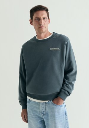 Hombre con cabello corto y oscuro, lleva una sudadera gris oscuro con logo y vaqueros azul claro, de pie con una mano en el bolsillo.